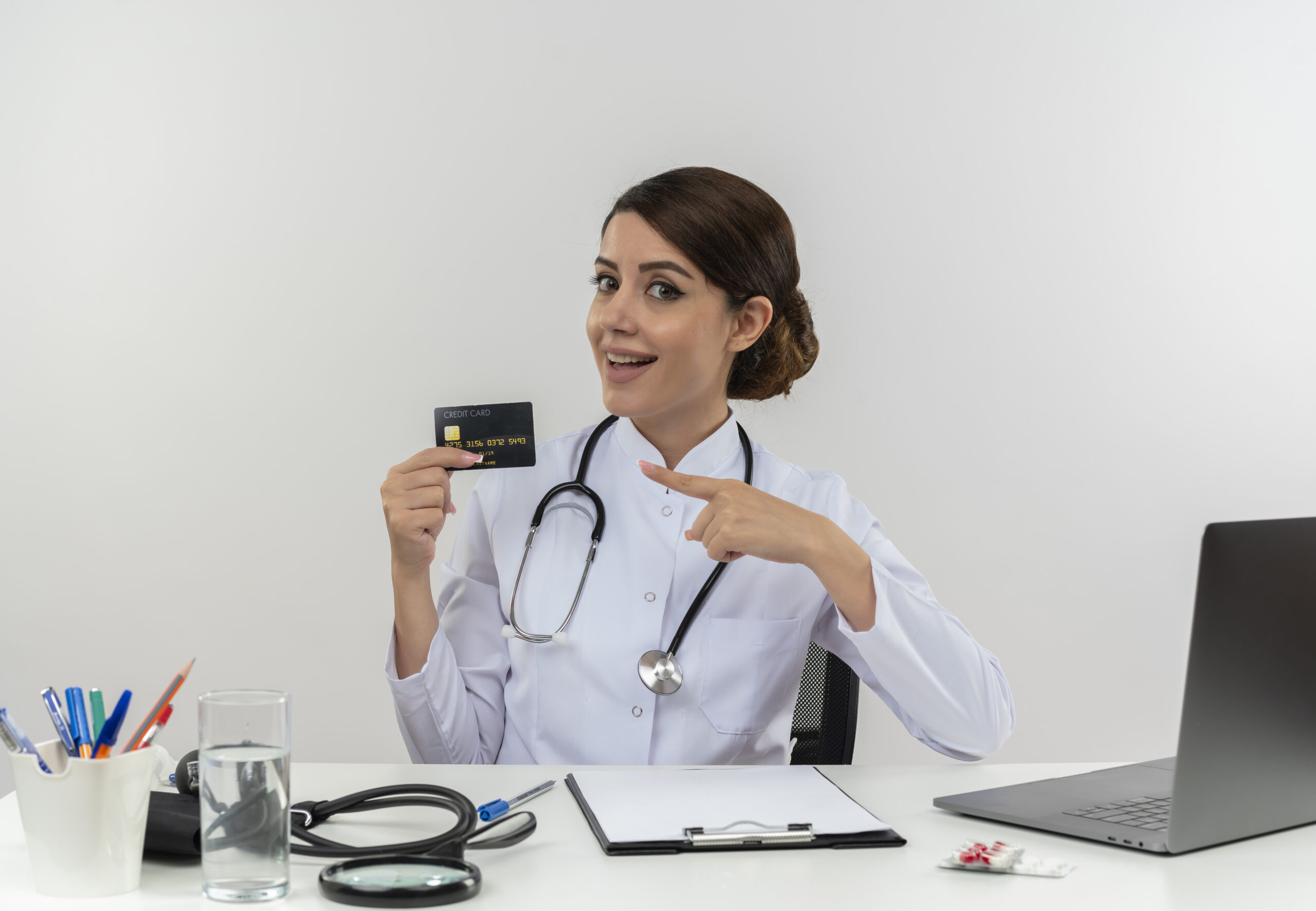 Home impressed young female doctor wearing medical robe and stethoscope sitting at desk with medical tools and laptop holding and pointing at credit card isolated on white background